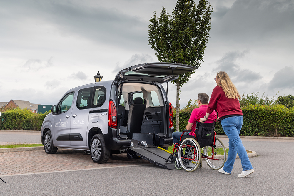 A woman pushes another woman in a wheelchair up a ramp into the back of an accessible car in a parking lot.